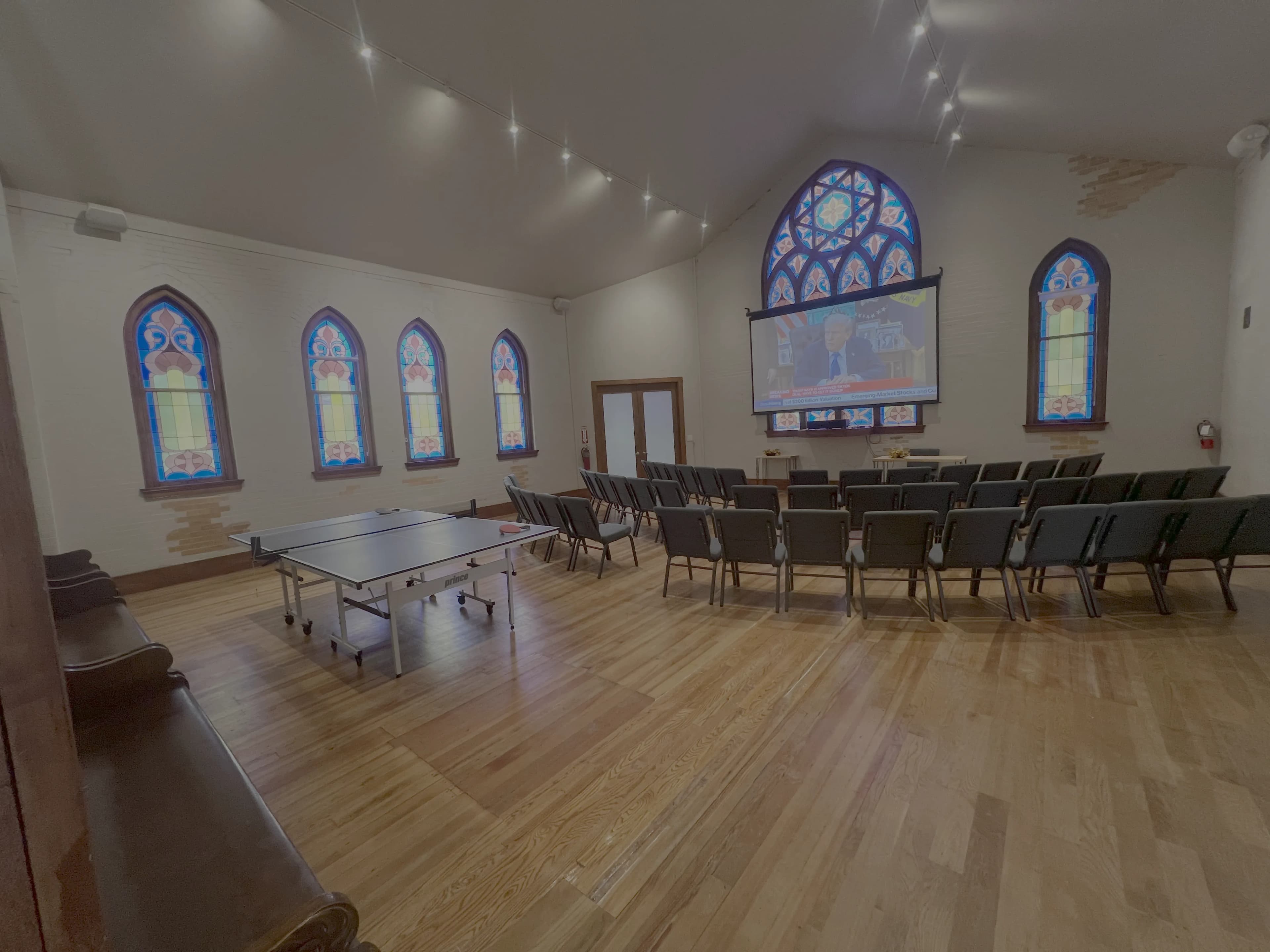 The restored 1905 hall at Merritt Workspace, with original stained glass, a hardwood floor, a projector screen, rows of seating and a ping-pong table, in Sloan's Lake, Denver